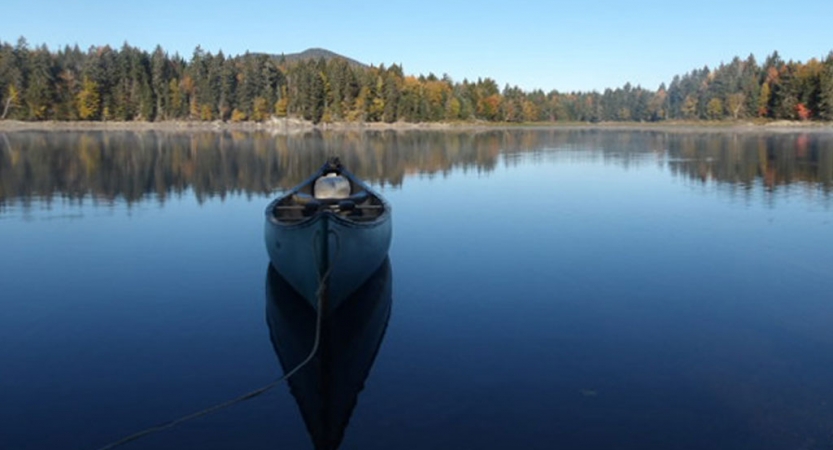 An empty canoe rests on very calm water, reflecting the blue sky and tree-lined shore in the background. 